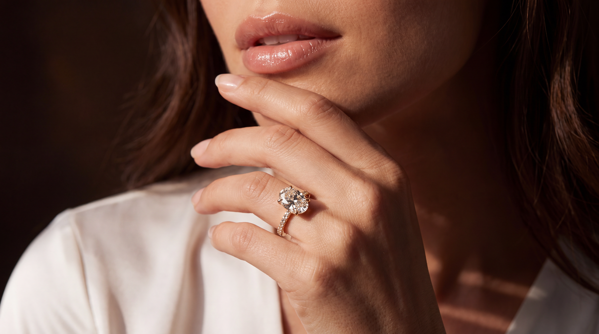 A close-up lifestyle shot of a woman wearing a rose gold ring with a round-cut clear gemstone on her finger, set against a dark background and warm lighting.