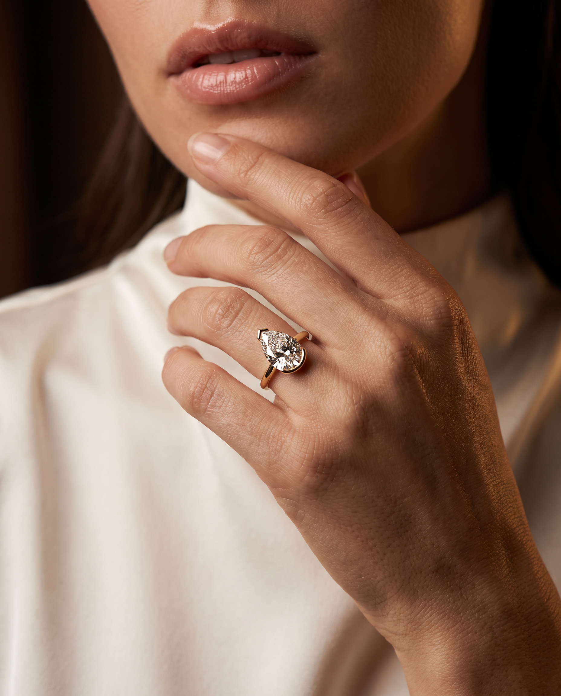 A close-up lifestyle shot of a model wearing a gold ring with a pear-cut clear lab diamond on her finger, dressed in a cream-colored high-neck top.