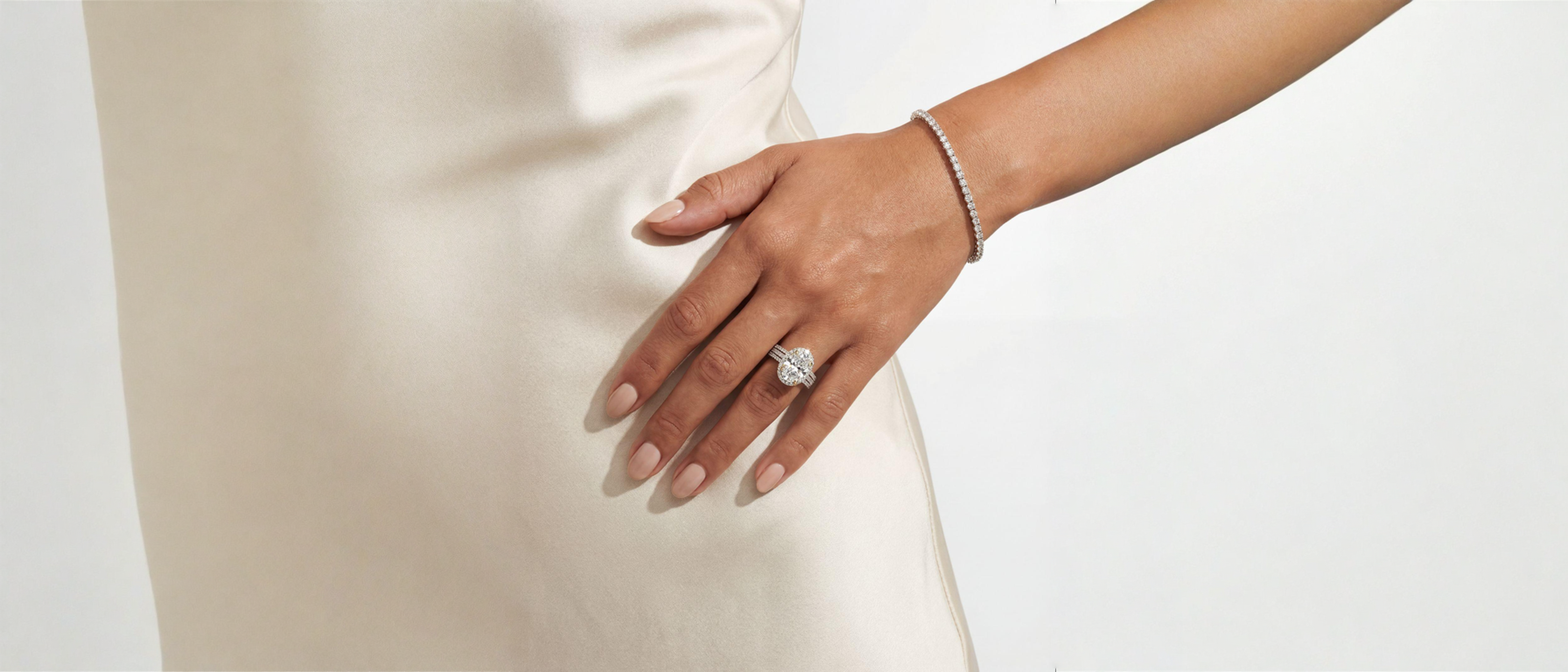 A close-up of a person's hand resting on a cream satin dress, showcasing a silver-toned diamond ring on the ring finger and a matching silver-toned beaded bracelet on the wrist.