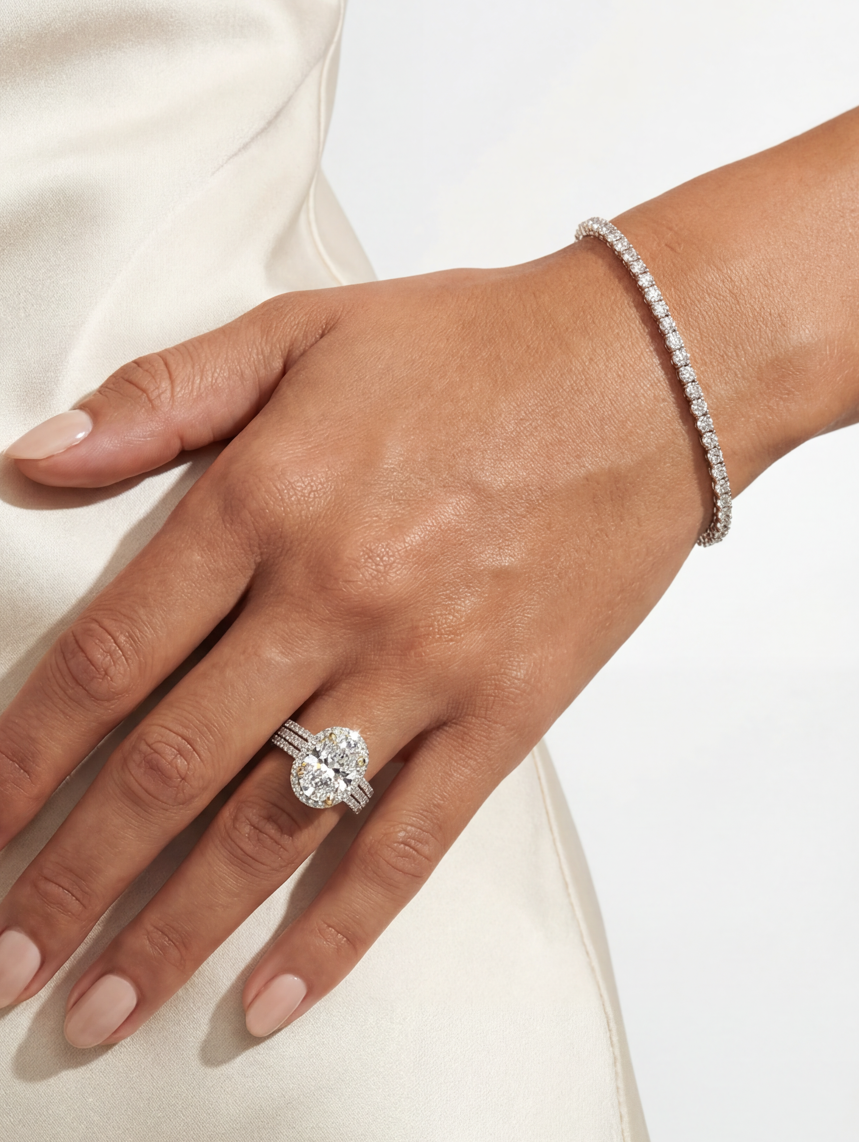 A close-up of a person's hand wearing a silver tennis bracelet and a matching ring with a large oval center stone, set against a soft cream satin fabric background.