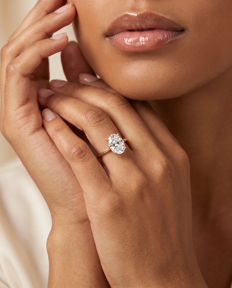 A close-up lifestyle shot of a model wearing a gold engagement ring with a round brilliant-cut diamond. The ring is shown on the model's ring finger, with her hands and lower face visible against a soft, warm beige background.
