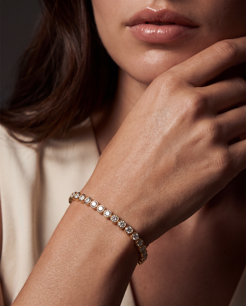 A close-up of a woman's hand and lower face wearing a delicate gold tennis bracelet with clear round-cut stones.
