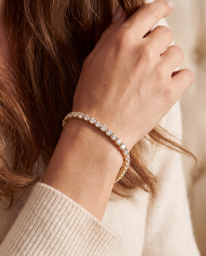 A close-up of a woman's hand and wrist wearing a delicate gold tennis bracelet adorned with clear stones, paired with a cream-colored knit sweater.
