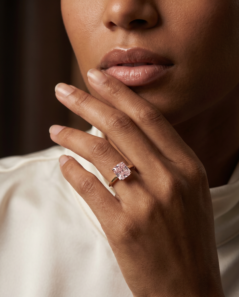 A close-up lifestyle shot of a model wearing a rose gold ring with a cushion-cut pink gemstone on her finger, with her hand resting near her chin.
