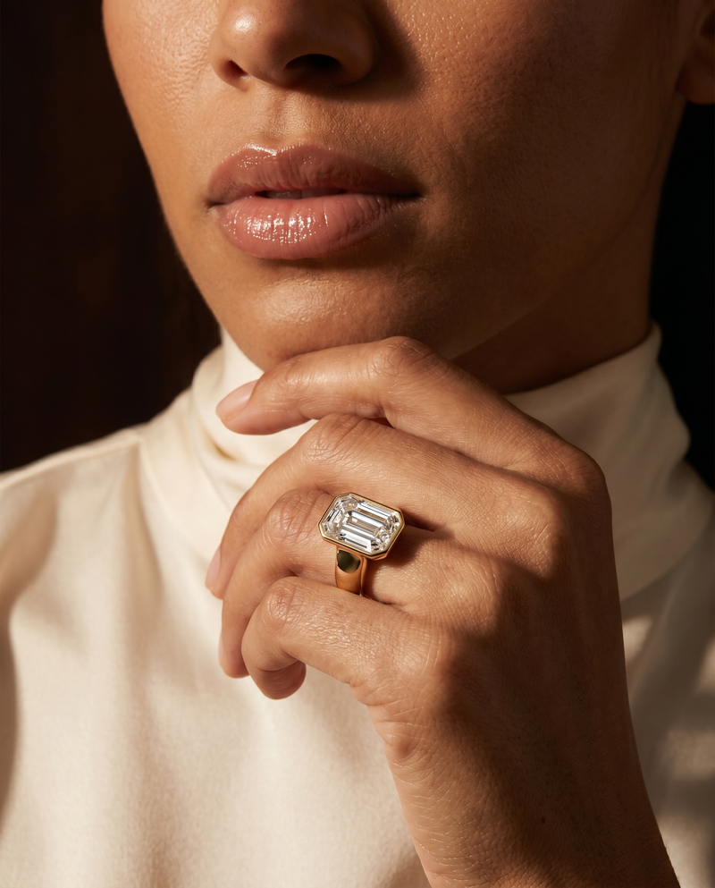 A close-up lifestyle shot of a model wearing a gold ring with a large rectangular emerald-cut clear gemstone, resting their chin on their hand.