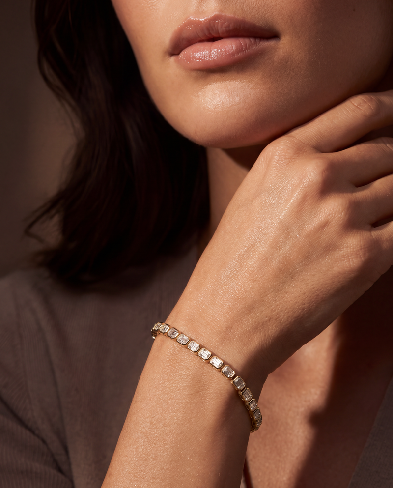 A close-up of a woman's hand and lower face wearing a delicate gold tennis bracelet with clear rectangular-cut stones, set against a warm, softly lit background.