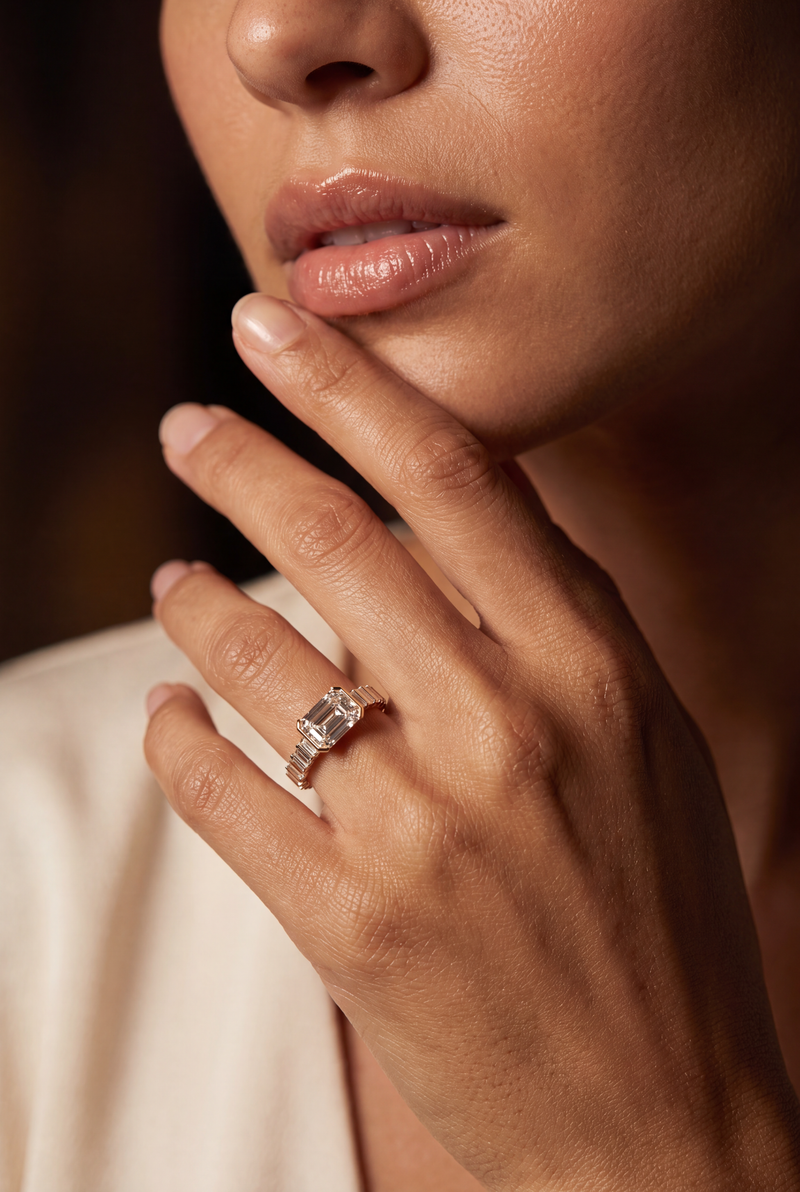 A close-up lifestyle shot of a model's hand resting near her chin, wearing a silver-toned ring with a rectangular clear gemstone.