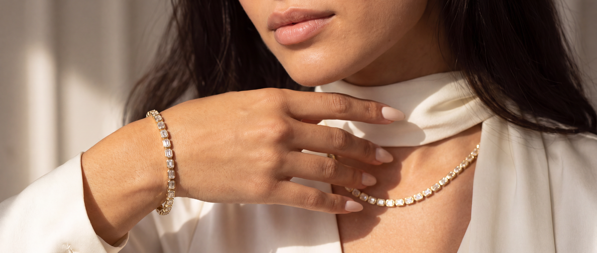 A close-up lifestyle shot of a woman wearing a delicate gold chain necklace and a matching gold bracelet, styled with a white high-neck top and draped fabric.