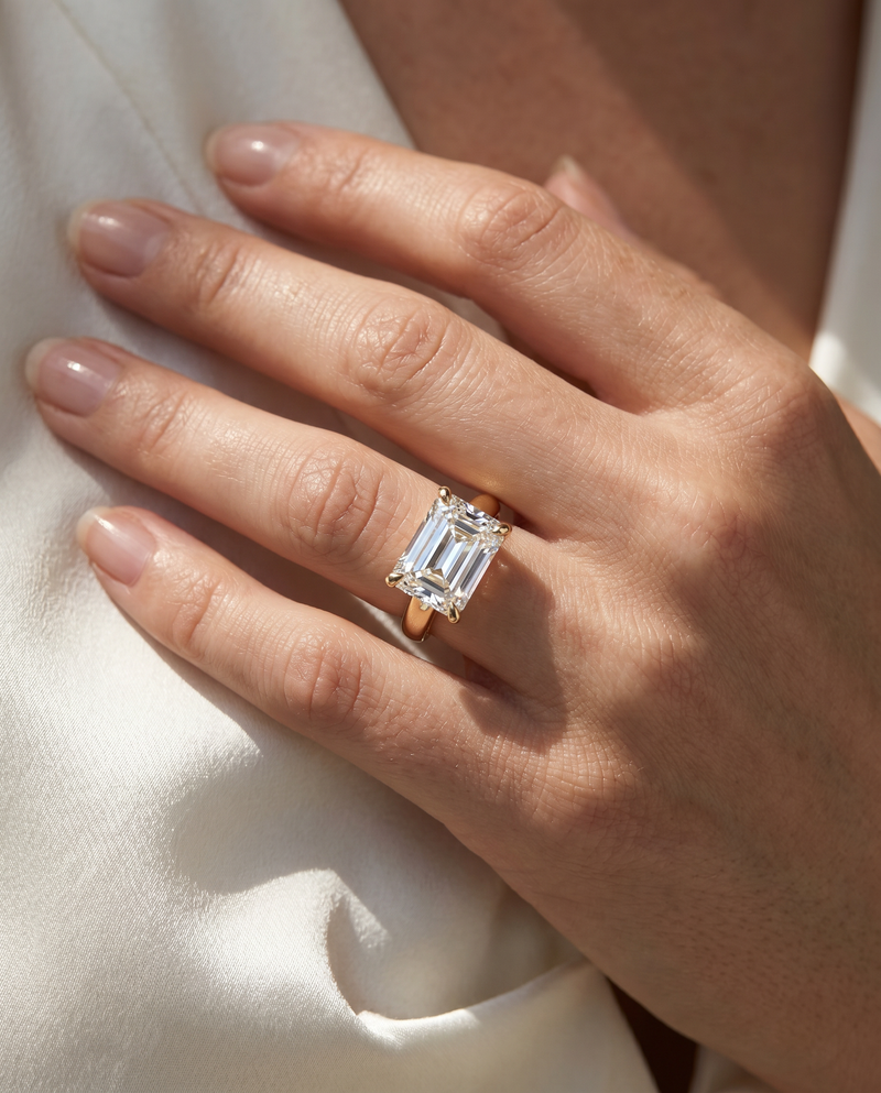 A close-up of a hand wearing a yellow gold ring with a large emerald-cut clear center stone, resting against white fabric.