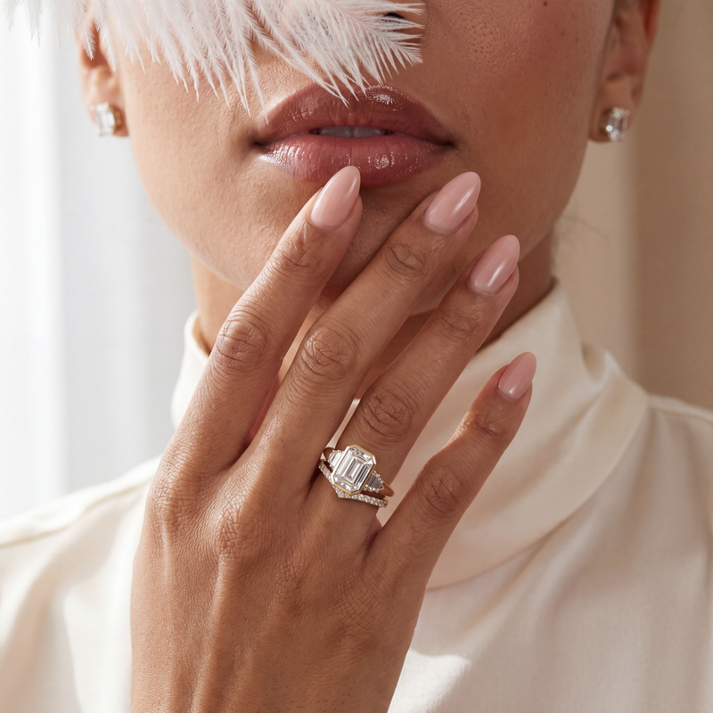 A close-up lifestyle shot of a model wearing a silver ring with a large emerald-cut clear center stone and smaller side stones. The model is wearing a cream-colored high-neck top, a white feather partially covers her face, and has manicured pink nails.