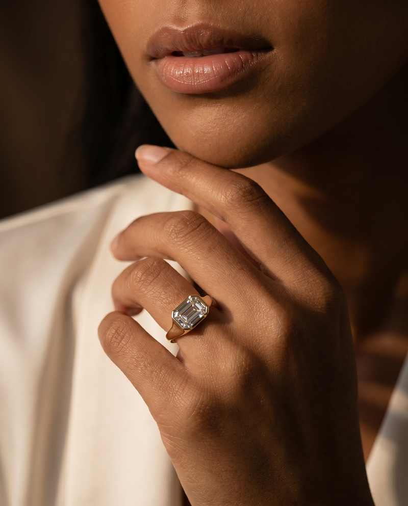 A close-up lifestyle shot of a model wearing a gold ring with a large rectangular clear gemstone on her finger, set against a dark background and white clothing.