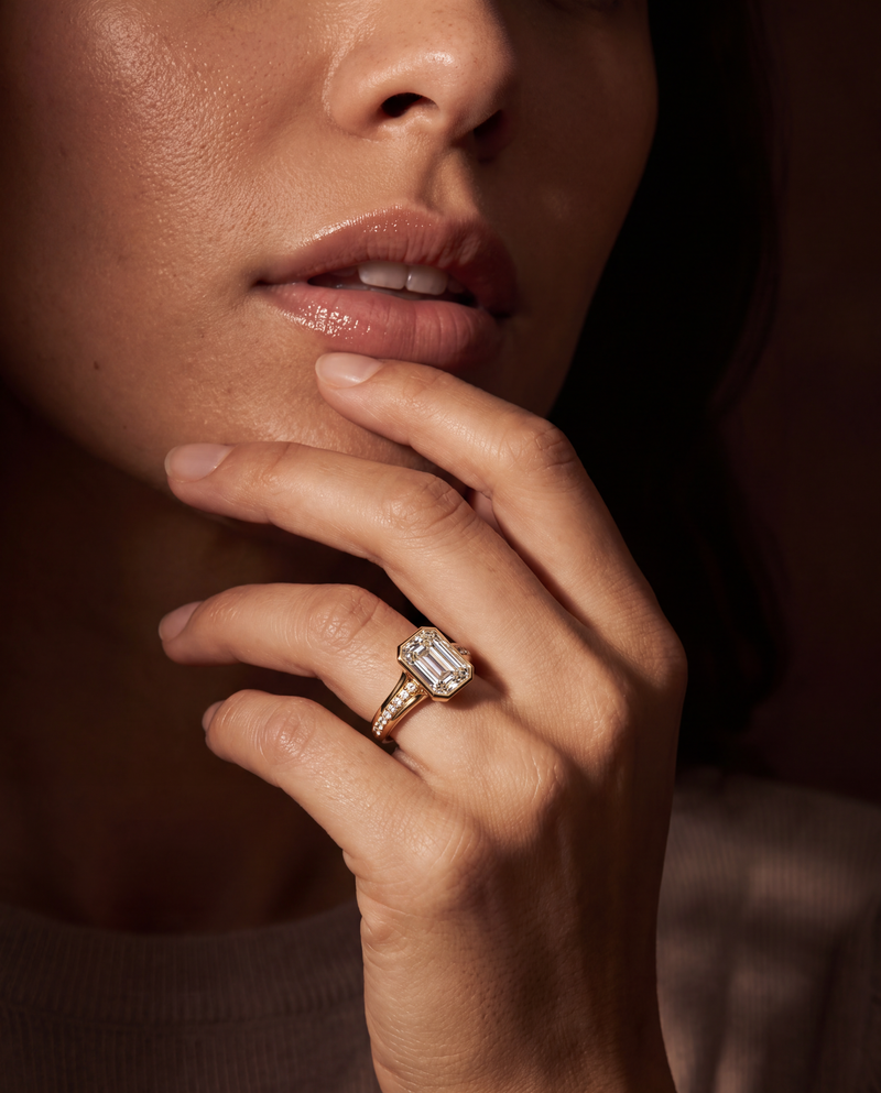 A close-up lifestyle shot of a model's hand resting near her chin, wearing a gold ring with a large rectangular emerald-cut diamond. The background is a very dark, warm-toned, shadowed setting.
