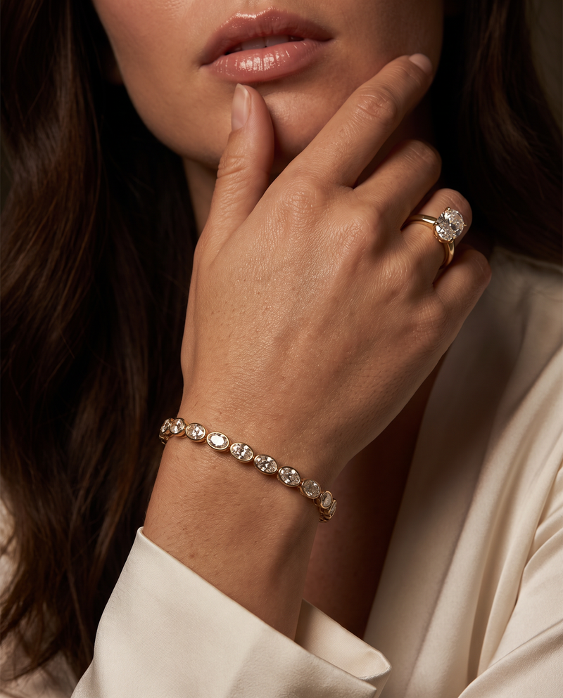 A close-up lifestyle shot of a woman wearing a delicate gold tennis bracelet with clear round-cut stones on her wrist, along with a gold ring on her finger, set against a dark background and cream-colored clothing.