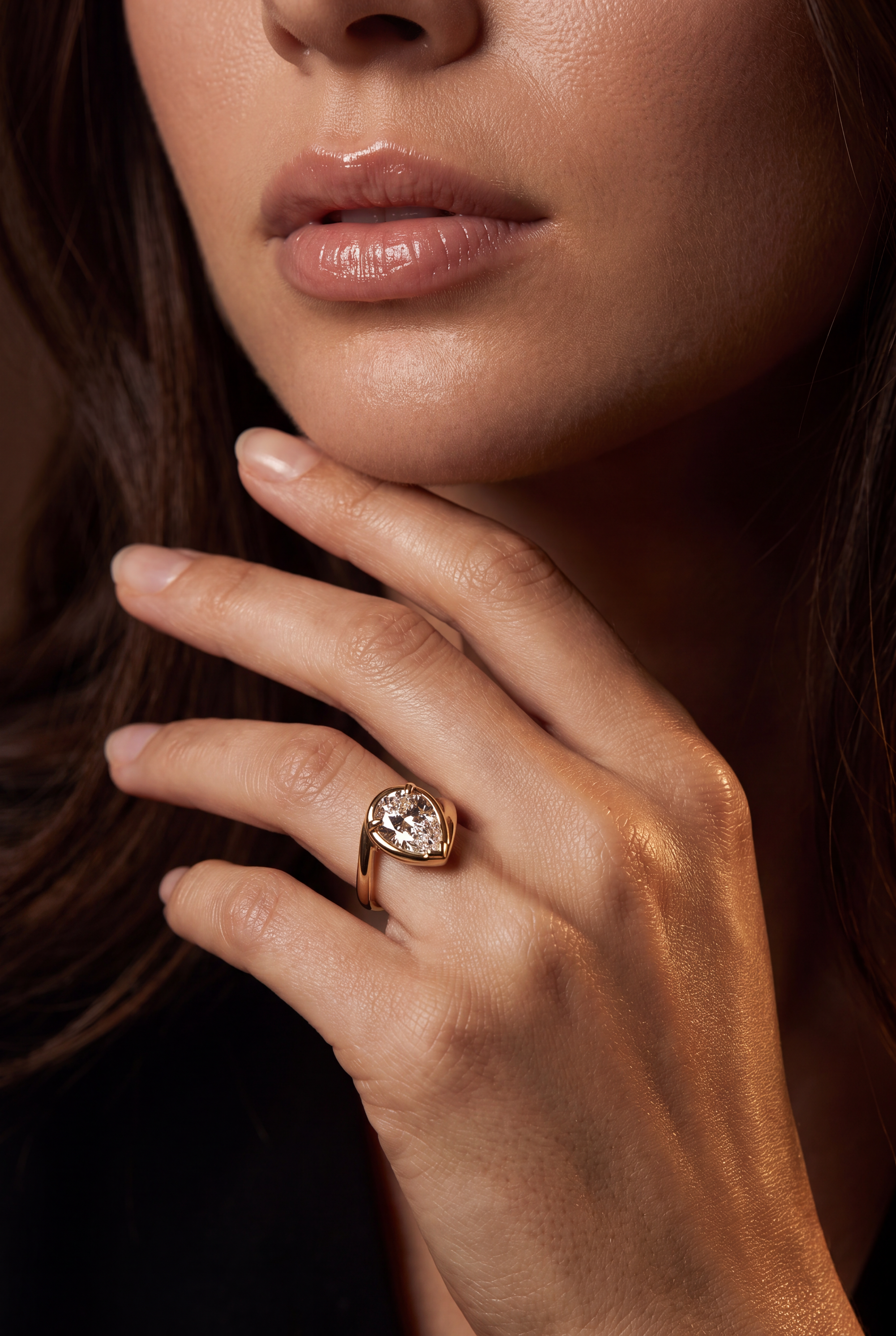 A close-up lifestyle shot of a model's hand wearing a gold ring with a large pear brilliant-cut diamond. The warm, golden-hour lighting highlights the skin and the metallic ring, set against a dark background.