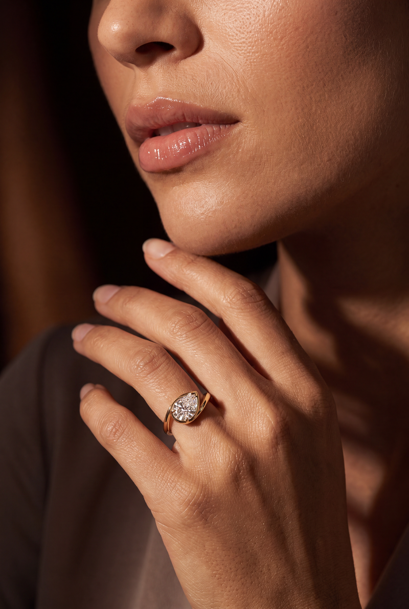 A close-up lifestyle shot of a woman's hand resting near her chin, wearing a gold ring with a marquise-cut clear gemstone. The image is shot in warm, dramatic lighting against a dark, blurred background.