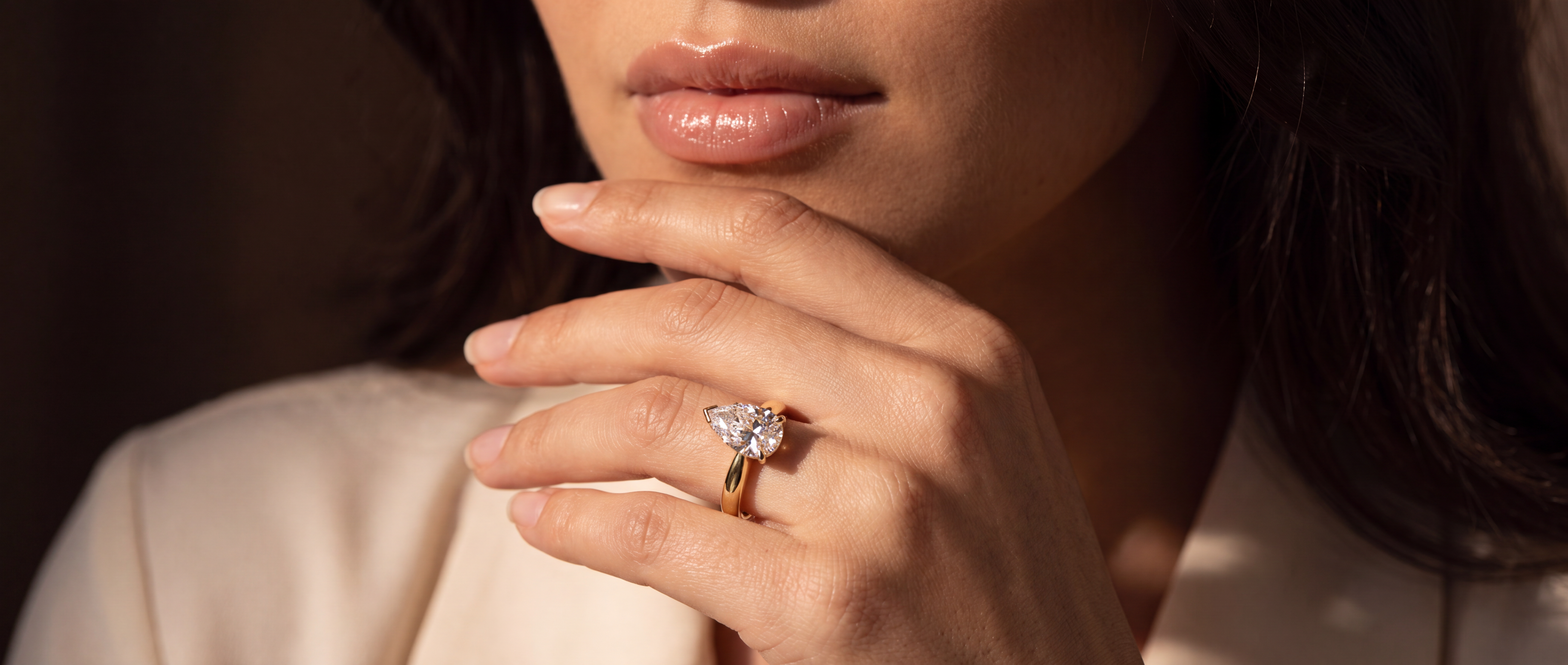 A close-up lifestyle shot of a woman wearing a yellow gold ring with a pear-shaped lab grown diamond on her finger, set against a dark background and a cream-colored blazer.