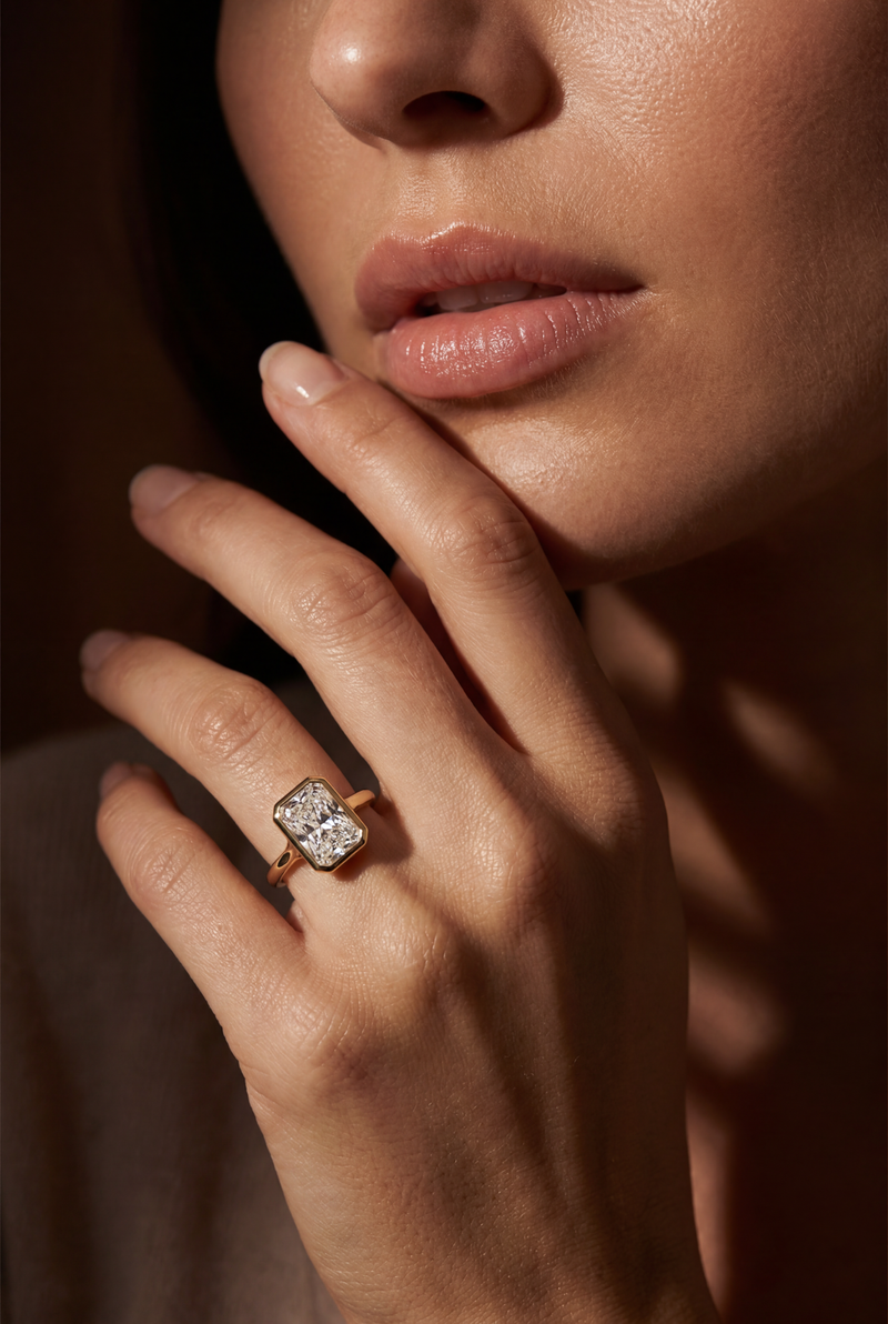 A close-up lifestyle shot of a model's hand wearing a rose gold ring with a rectangular cushion-cut clear gemstone, set against a dark, softly lit background.