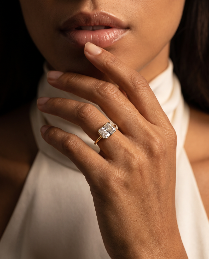 A close-up lifestyle shot of a model wearing a gold ring with a rectangular cushion-cut clear gemstone. The ring is shown on her ring finger, with her hand resting near her chin against a dark background and a white high-neck top.