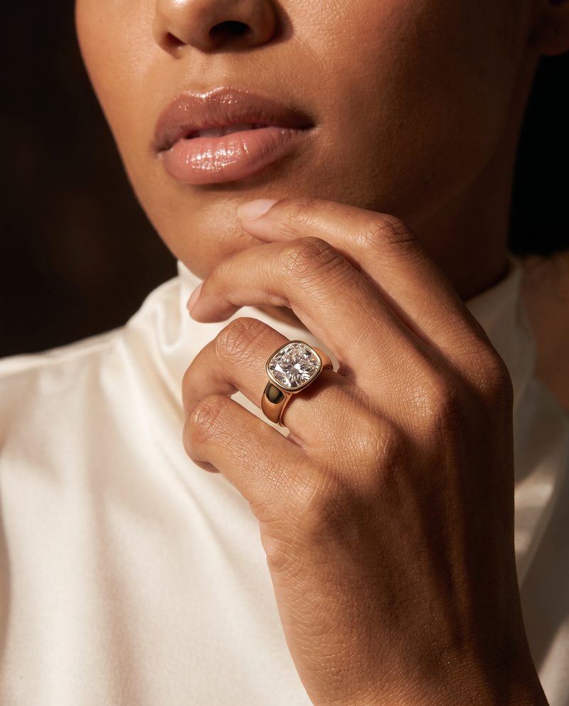 A close-up lifestyle shot of a model wearing a cushion-cut diamond ring with a gold band and a small black accent on the side, set against a dark background and white satin shirt.