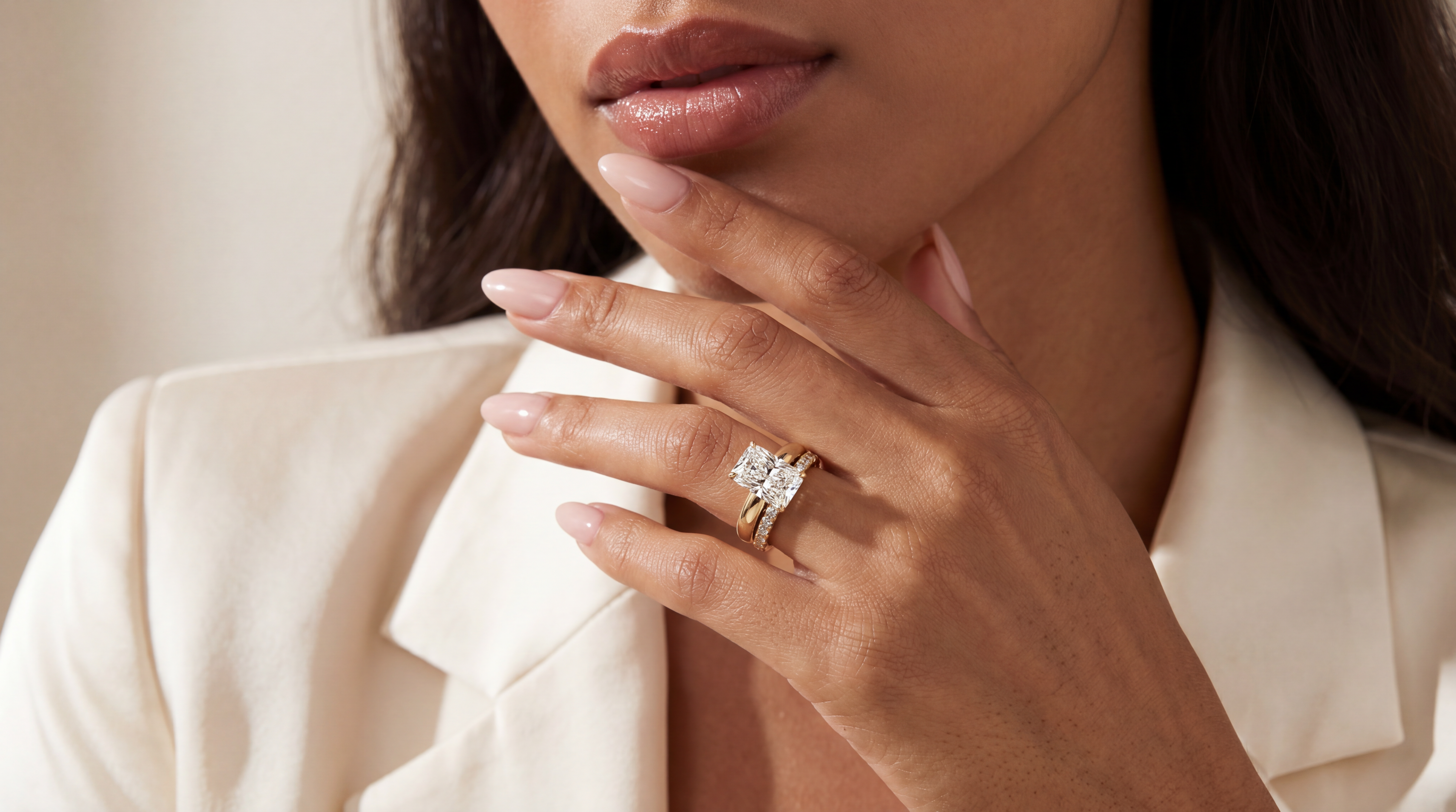 A close-up lifestyle shot of a woman wearing a yellow gold ring with a large round-cut clear gemstone and a matching gold band, shown on her hand against a soft beige background.