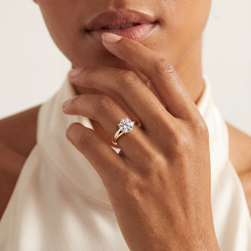 A close-up lifestyle shot of a model wearing a yellow gold engagement ring with a round brilliant-cut clear gemstone on her finger. The background is a soft, warm off-white.