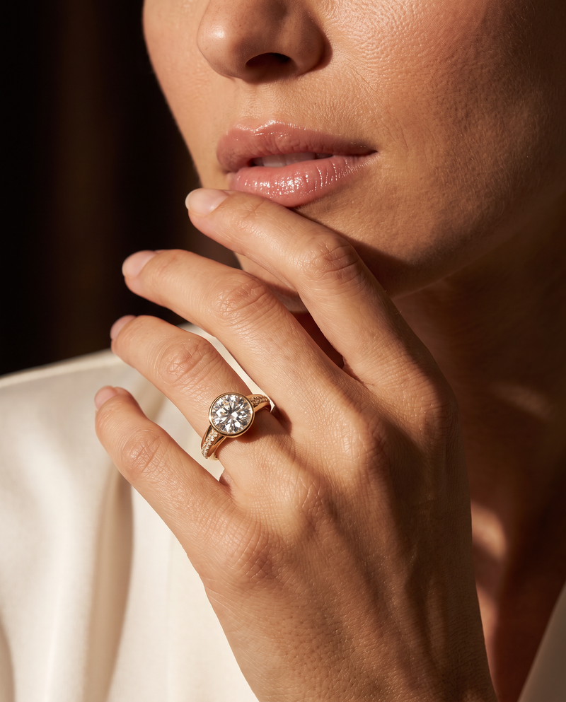 A close-up lifestyle shot of a woman wearing a gold ring with a round-cut clear gemstone on her finger, set against a dark background and warm light.