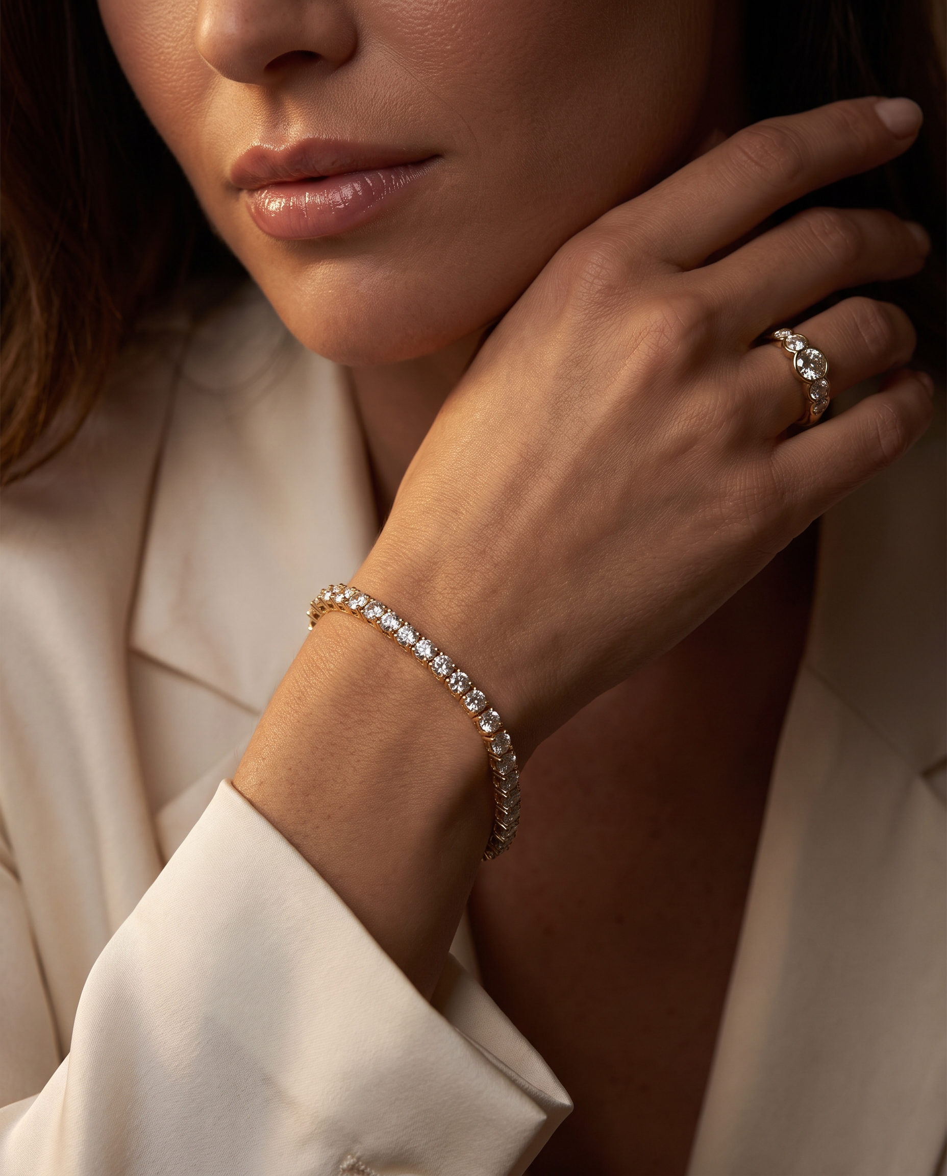 A close-up lifestyle shot of a model wearing a delicate gold tennis bracelet on her wrist and a matching gold ring on her finger, paired with a cream-colored blazer.
