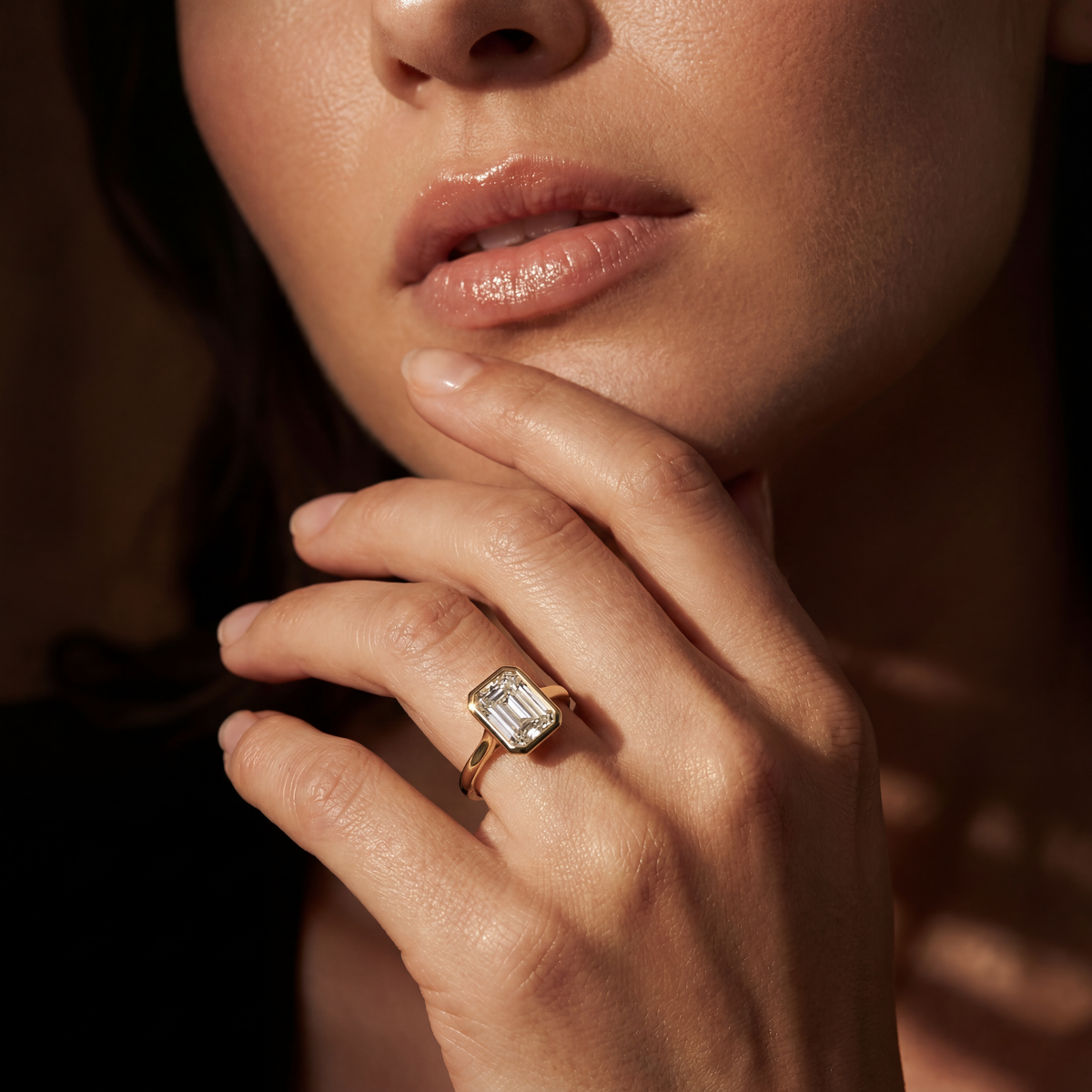 A close-up lifestyle shot of a woman's hand resting near her chin, wearing a gold ring with a large rectangular emerald-cut clear gemstone.