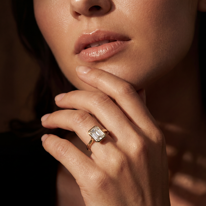 A close-up lifestyle shot of a woman's hand resting near her chin, wearing a gold ring with a large rectangular emerald-cut clear gemstone.