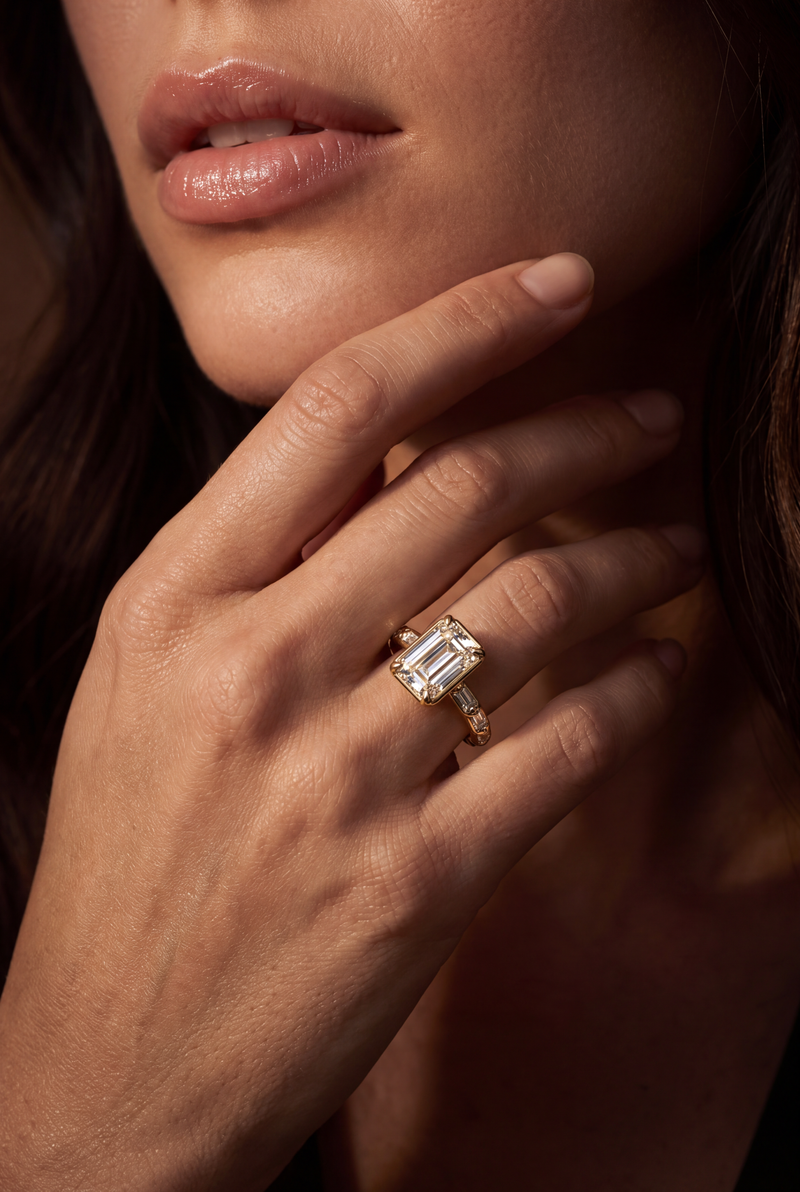 A close-up lifestyle shot of a model's hand resting near her chin, wearing a yellow gold ring with a large rectangular emerald-cut diamond. The diamond is clear and does not contribute to the color mapping according to the schema rules.