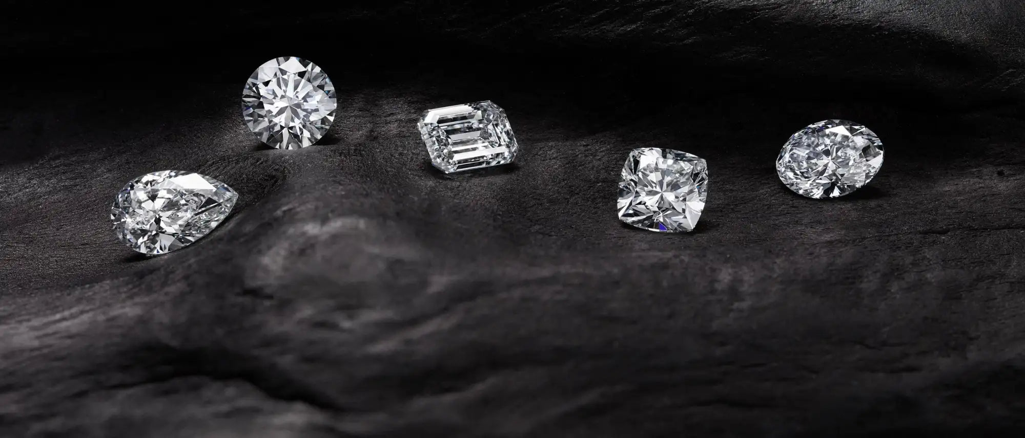 Five sparkling, clear-cut diamonds of varying shapes—round, emerald, cushion, pear and oval—are arranged on a dark, textured surface.